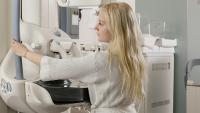 Womaan in robe stands at machine during mammogram while radiologist operates imaging equipment.